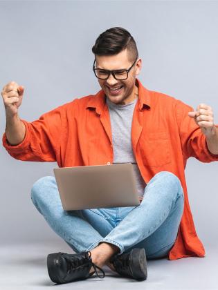 successful-winner-portrait-happy-young-man-sitting-floor-using-laptop-celebrating-success-victory-isolated-white-grey-background